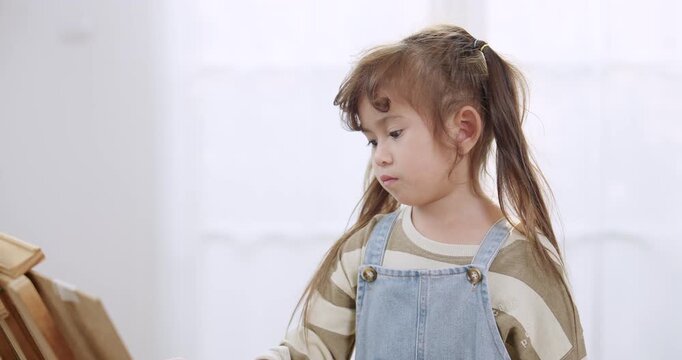 Preschool asian daughter stands near easel canvas, mixing color on brush during creative playtime, quiet homeschool art practice, fine motor skill training, imagination growth, bright indoor room.