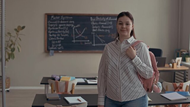 Medium eye level portrait of Caucasian female student smiling at camera with backpack on shoulder, standing in classroom with economics theory on blackboard in background