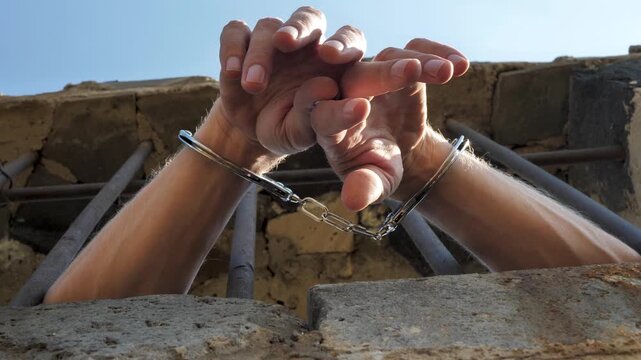 Low angle view: male hands in handcuffs sticking out of prison window through bars. Handcuffed person outside window to prevent escape. Convicted hands with metal bracelets. Concept of justice