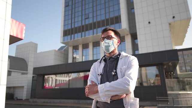 Confident doctor wearing medical mask posing outside modern hospital while camera moves in semicircle around him. Concept of healthcare, professionalism and safety in urban environment.