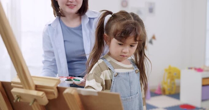 Preschool asian daughter reaches easel canvas, dips brush into paint palette while single mother supervises, home education concept, calm bonding, creative learning inside playroom, fine motor skill.