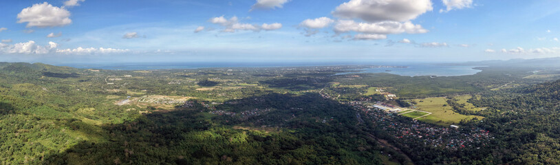 Fototapeta premium Wide aerial panorama from Irawan mountains shows the valley town spreading toward Honda Bay and open ocean with lush green hills in foreground and bright blue sky stretching across Palawan horizon