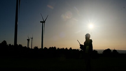 Engineer silhouette operating a drone controller at a wind farm during sunset, checking renewable energy production and sustainable technology infrastructure © DawDunia