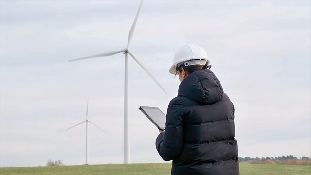 Engineer in hard hat checking wind turbine operation using a digital tablet at a wind farm, managing clean, renewable energy production and sustainability efforts