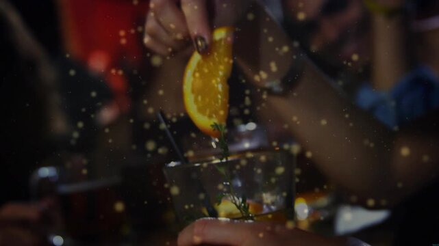 Man holding lowball at bar, woman lowering, pressing orange wedge into drink for aroma, golden glow