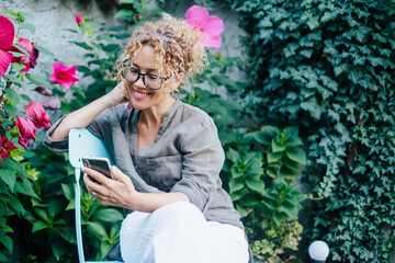 Smiling woman relaxing in a lush garden chair while checking her mobile phone and enjoying the...