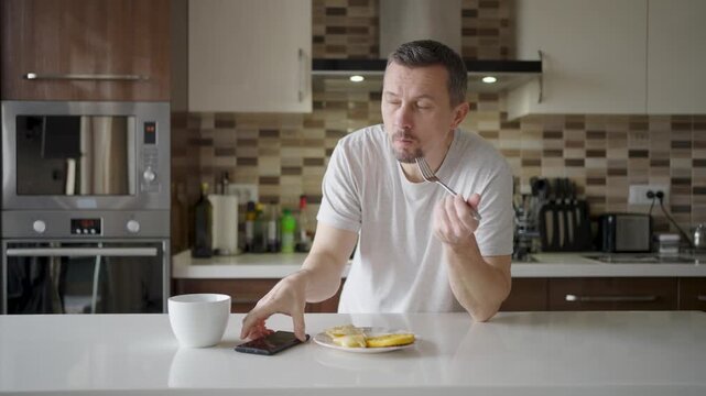 Man using smartphone while having breakfast in kitchen. A man in a t-shirt eating pancakes and scrolling on his phone at a white kitchen island. Concept of morning routine, news, and digital lifestyle