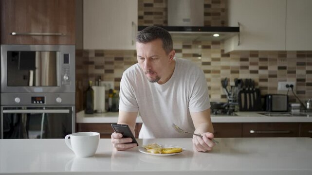 Man using smartphone while having breakfast in kitchen. A man in a t-shirt eating pancakes and scrolling on his phone at a white kitchen island. Concept of morning routine, news, and digital lifestyle