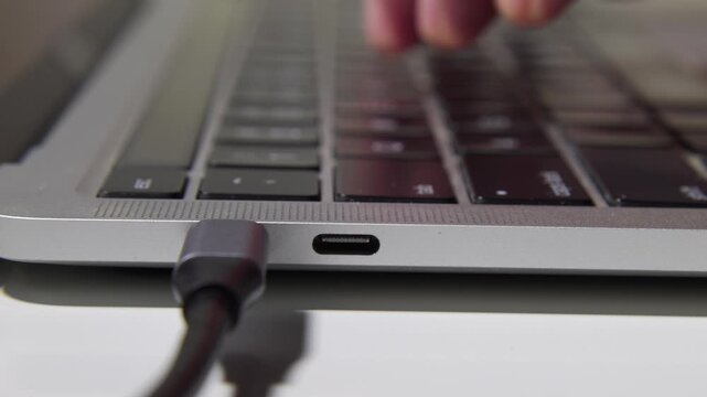 Macro shot of a hand connecting a USB-C power cable into a laptop port, followed by fingers starting to type on the keyboard. Concept of starting work, productivity, and technology integration.