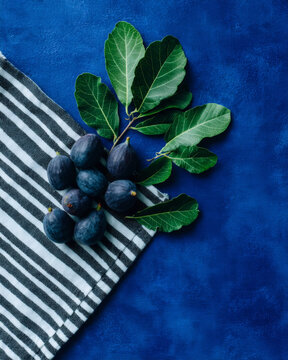 Top view flat lay of fresh dark figs with green leaves on striped linen napkin and textured navy blue background, gourmet food photography with copy space.