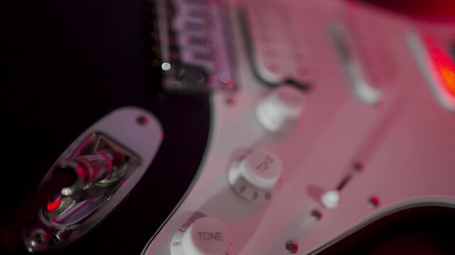 Close-up of Musician Plugging Jack Cable into Electric Guitar. A musician hand connects a silver jack cable into a black electric guitar with white pickguard under moody red stage lighting