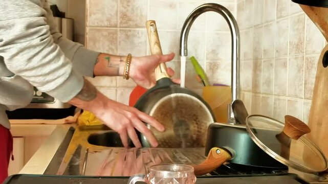 A man, whose arms are only framed, cleans a pan by rubbing it with a sponge. The man uses the kitchen sink as a support point