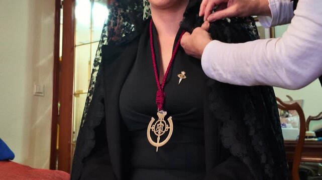A woman being helped to adjust her traditional Spanish mourning dress featuring a black lace mantilla veil and a religious golden medal on a red cord during Holy Week