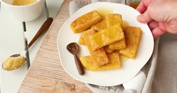 Crispy fried polenta sticks on plate with parchment paper. Hand using fork to pick golden cornmeal appetizer served on rustic wooden board with garlic and dipping sauce.