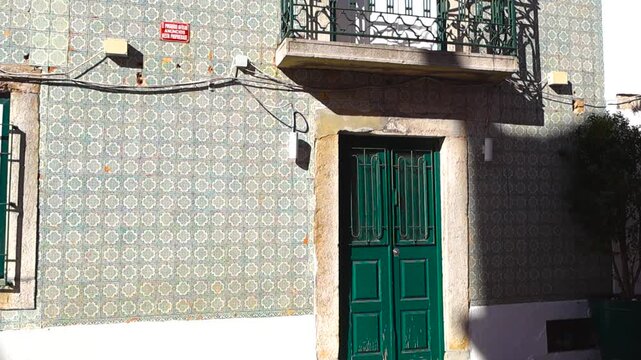Authentic facade of a traditional Portuguese building in Lisbon, featuring intricate Azulejo ceramic tiles, a classic green wooden door, and an ornate balcony with moving sunlight and shadow