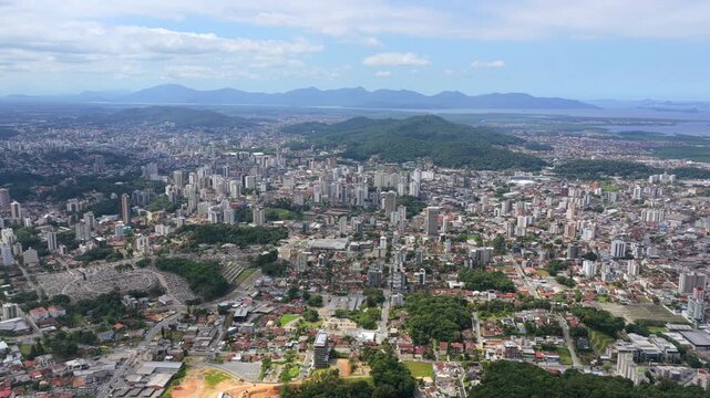 Wide aerial panorama of Joinville city with dense skyline, green hills, and coastal landscape.