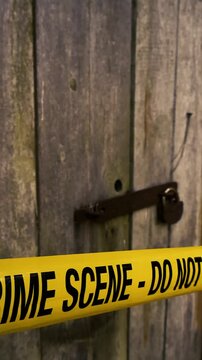 Yellow police tape with Crime Scene Do Not Enter warning text blocking access to an old wooden door, secured with a rusty hasp and padlock, creating a suspenseful atmosphere