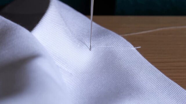 A close-up video shows a sewing needle rhythmically piercing white fabric, highlighting the precision and delicate texture of the stitching process against a wooden backdrop.