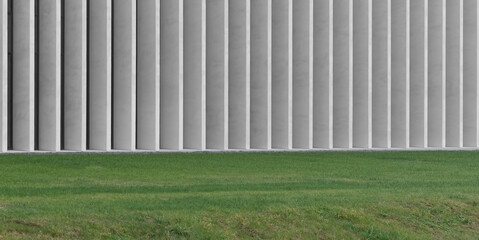 Fototapeta premium Concrete column wall runs along green grass creating contrast between structure and nature. Repeating vertical elements form strong linear rhythm across facade. Concept of minimal architecture,