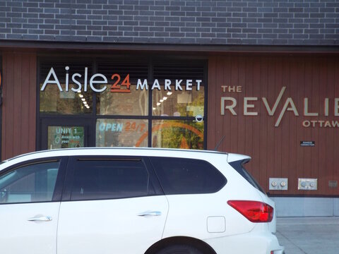 Ottawa, Ontario, Canada &ndash; July 26, 2025: The "Aisle 24 Market "storefront features modern signage, wood-style paneling at The Revalie, and a white SUV parked in front at 770 Brookfield Rd.
