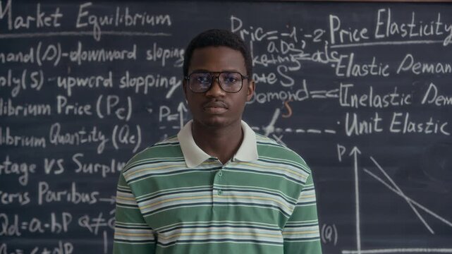 Medium eye level portrait of African American male student standing in classroom with chalkboard full of economic theory and graphs in background