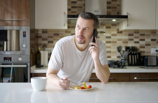 A man talking on his mobile phone and smiling while eating pancakes in a modern kitchen. Concept of morning call, communication, and multitasking at home