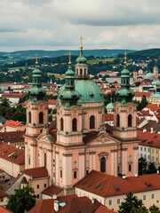 Obraz premium High-Angle View of a Grand Baroque Church with Green Domes and Golden Crosses in a Historic European Town, Surrounded by Red-Roofed Buildings and Hills under a Cloudy Sky