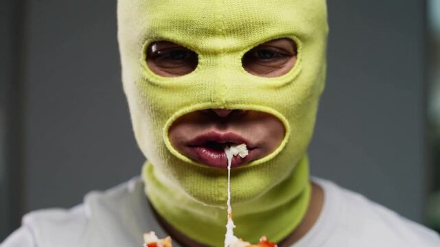 Hunger leading masked man wearing neon yellow balaclava biting pizza slice closeup, pulling cheese
