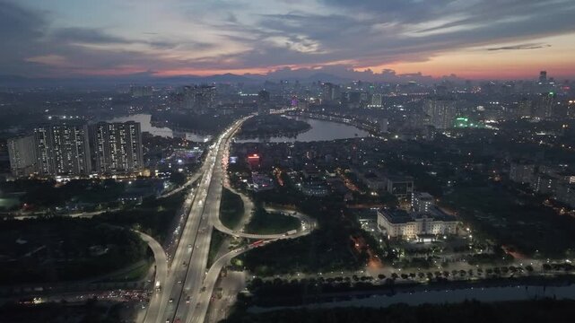 Nighttime drone panorama of major highway interchange in Hanoi with bright streetlights, flowing traffic, and dense urban skyline in background