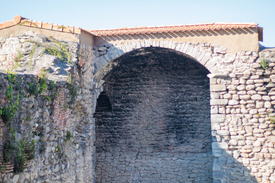 Detalle de una estructura de piedra antigua con un gran arco de medio punto y tejado de tejas. La mamposter&iacute;a hist&oacute;rica muestra texturas rugosas y vegetaci&oacute;n silvestre creciendo entre las grietas.