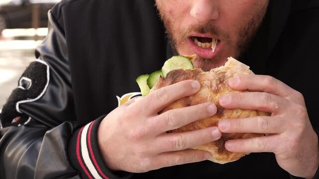 A close-up, high-angle shot focusing on a young man with a beard as he takes a large bite of a fresh doner kebab or falafel sandwich. The sandwich is filled with cucumbers and meat in a toasted pita. 