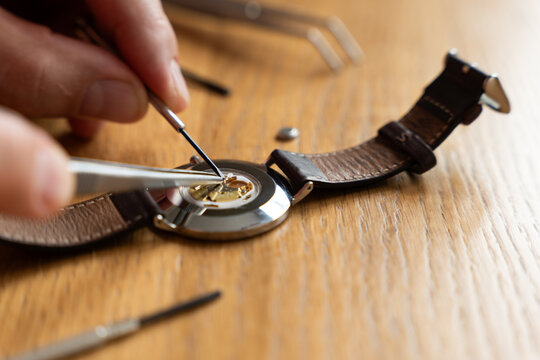 Individual carefully repairs watch movement using tweezers on a wooden workbench, surrounded by various watch parts and tools