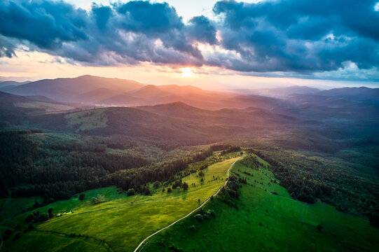 Aerial view breathtaking sunset over forest. Sun's rays break through dramatic clouds, casting golden glow across rolling hills and valleys. Distant mountains creating serene and picturesque scene.