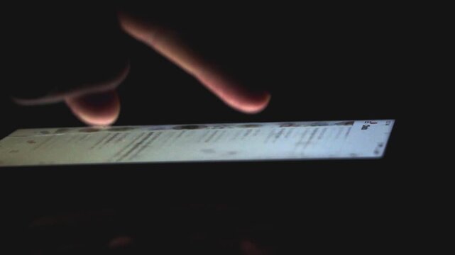 Dramatic and moody video footage captures a close-up, side-angle view of a person's finger actively scrolling and swiping on a brightly glowing smartphone screen