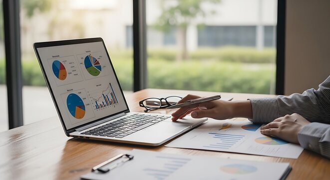 Close-up of hands working on a laptop with financial charts & reports on a wooden table