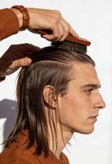 Naklejka premium Vertical photo of a young man with long wet hair being styled. Professional grooming with a wooden brush. Side profile of male model in brown jacket against white background