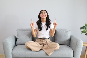 A smiling young Asian woman looks and points up at the empty space for your ad text, sitting cross-legged on the sofa in the living room of the house. © ArvStd