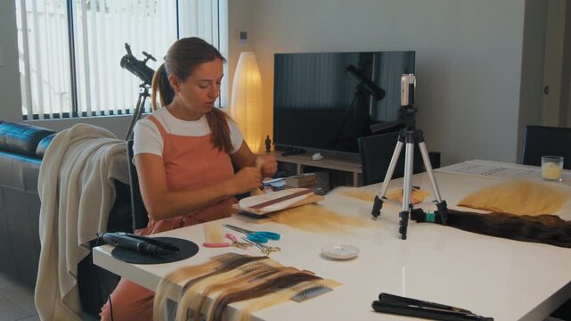 A woman is working on hair extensions at a table in a living room with a mobile phone recording her. Person doing online training in hair extensions