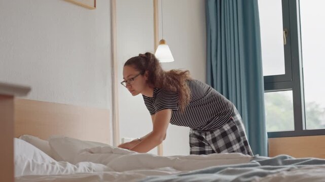 Woman making hotel bed in bright modern room, adjusting sheets and pillows, sunlight through window, blue curtains, bedside lamp, focused on neatness, solo traveler preparing space for stay