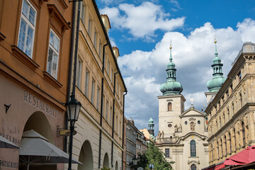 Obraz premium Historic Baroque Church Building With Green Twin Spires Dominating An Old Town Street Scene Lined With Traditional Prague Architecture On A Sunny Day, Czech Republic