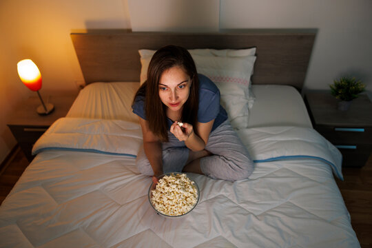 Woman eating popcorn while watching funny movie on TV in bed at home at night