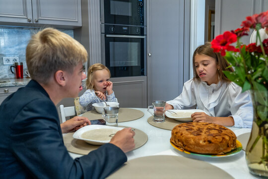 Candid family mealtime scene with three children around modern kitchen table, featuring displeased girl, nose-picking toddler, and large delicious pie, capturing everyday reality of family life.