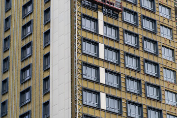Modern high-rise building facade under construction, showcasing multiple windows, exposed thermal insulation, and installation of contemporary exterior cladding panels for urban development. © sommersby