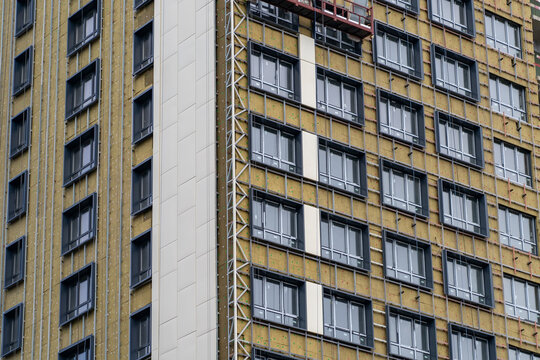 Modern high-rise building facade under construction, showcasing multiple windows, exposed thermal insulation, and installation of contemporary exterior cladding panels for urban development.