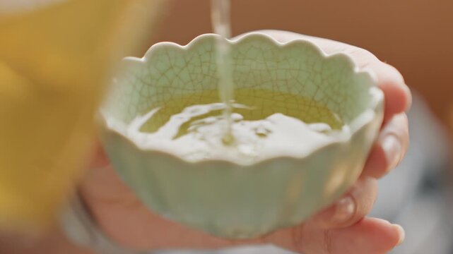 Close shot of hand pouring tea into scalloped celadon cup, warm golden liquid, soft blurred background, subtle steam rising, ceramic glaze crackle visible, hands offering hospitality, cozy intimate