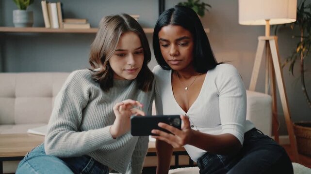 Holding smartphone right woman showing pal tapping phone on sofa viewing content wearing white top