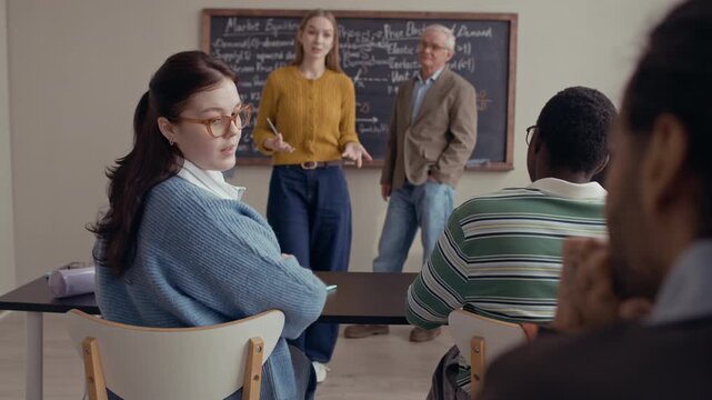 Over shoulder medium shot of two students speaking while their classmate standing at chalkboard with senior male professor giving presentation during economics lesson