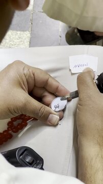 Detailed close-up of an artisan using a rotary tool to engrave &ldquo;Afreen&rdquo; on a white charm of red prayer beads, showcasing fine craftsmanship and traditional religious souvenir making.