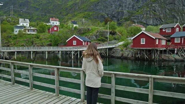 Woman observing red fishing cabins across a calm harbour in Lofoten, Norway. Peaceful coastal village with reflections and mountains.