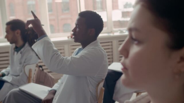 Closeup of female medical student nodding while listening and her African American male classmate raising hand to participate in discussion during interactive healthcare lesson in modern classroom